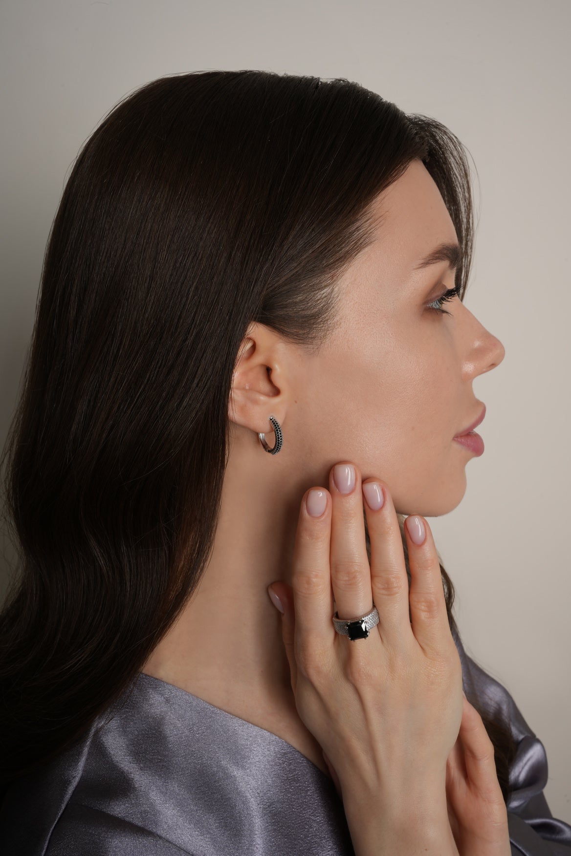 Woman wearing earrings and a ring, touching her neck against a neutral background