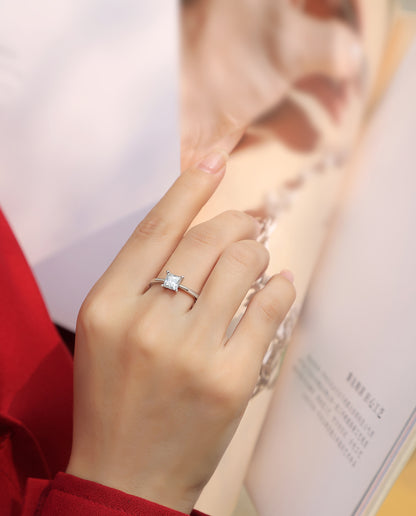 Elegant moissanite solitaire ring by Olivia Star Jewelry, princess cut stone set in 925 sterling silver, shown on hand in lifestyle photo with fashion magazine